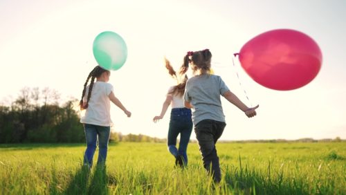 Crianças brincando com balões coloridos em um campo aberto iluminado pelo sol
