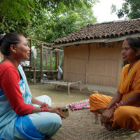 Duas mulheres sentadas em posição de lótus, uma em frente a outra, conversam em frente a uma casa simples, em ambiente arborizado