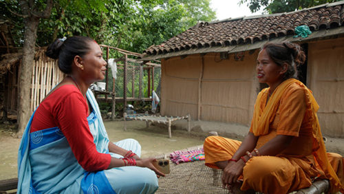 Duas mulheres sentadas em posição de lótus, uma em frente a outra, conversam em frente a uma casa simples, em ambiente arborizado