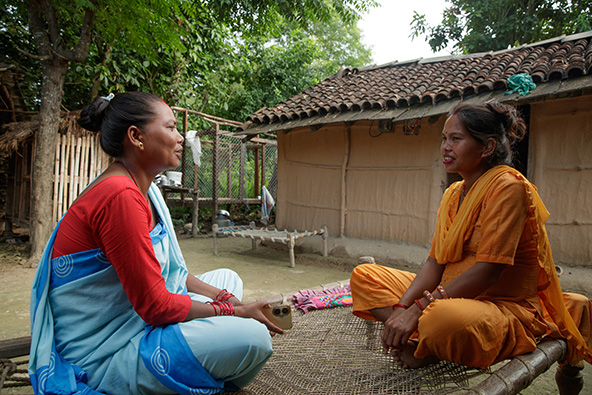 Duas mulheres sentadas em posição de lótus, uma em frente a outra, conversam em frente a uma casa simples, em ambiente arborizado