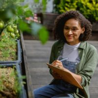 foto de uma mulher sorrindo e portanto um bloco de notas, onde aparenta fazer notas sobre plantas que estão ao seu redor
