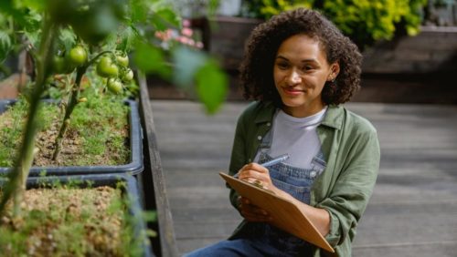 foto de uma mulher sorrindo e portanto um bloco de notas, onde aparenta fazer notas sobre plantas que estão ao seu redor