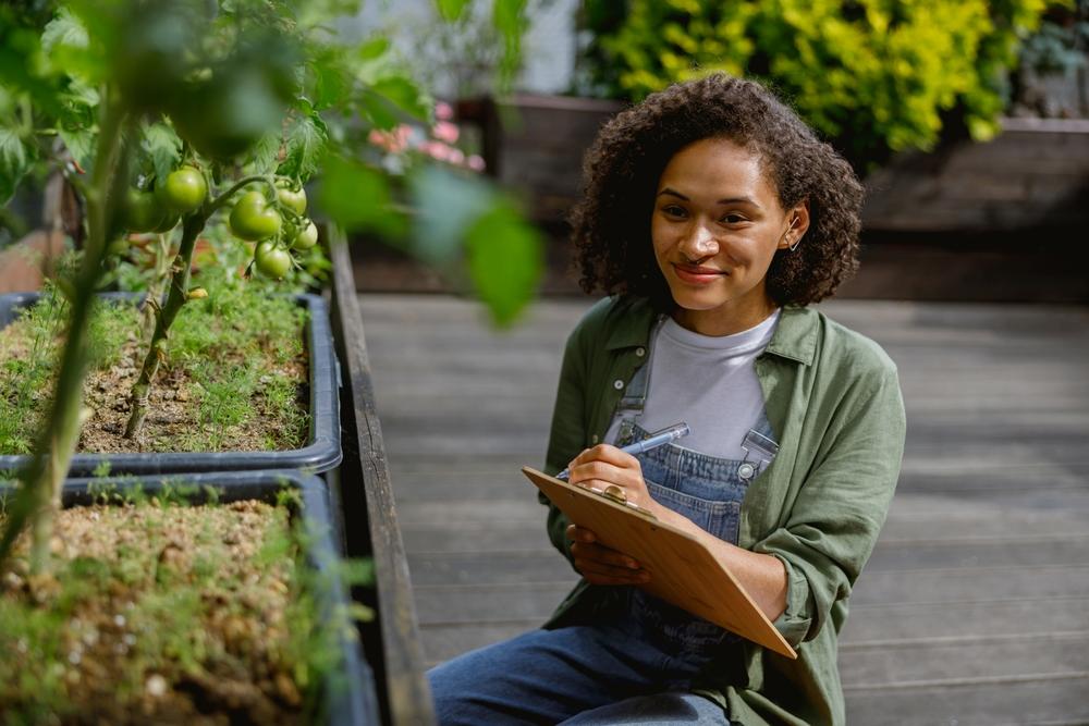 foto de uma mulher sorrindo e portanto um bloco de notas, onde aparenta fazer notas sobre plantas que estão ao seu redor