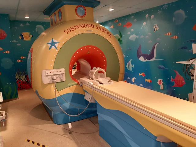 foto de uma máquina para realização de tomografia em sala temática com pinturas na parede de peixes e oceano. Na máquina, também decorada, está escrito: "Submarino Sabará"