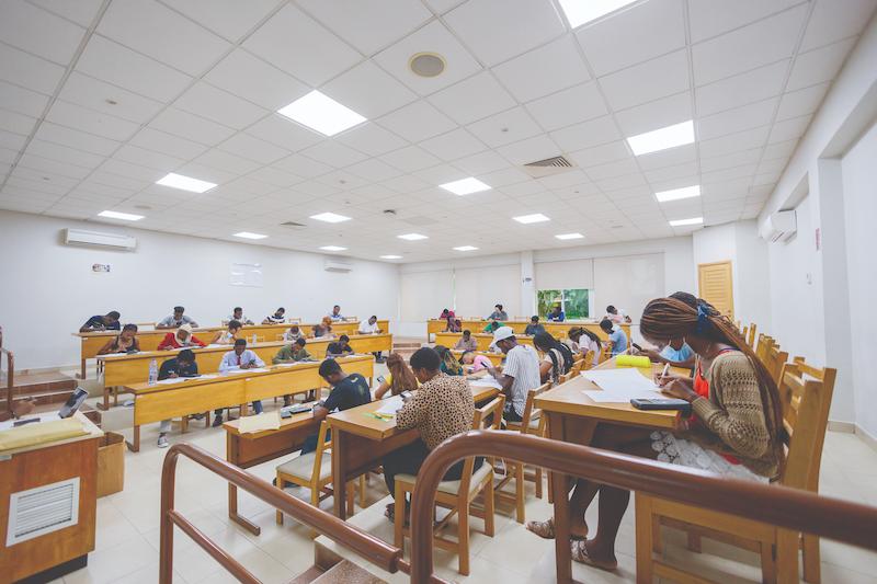 Imagem de uma sala de aula com paredes brancas, bancos e mesas de madeira, onde alunos de debruçam sobre papéis de estudo
