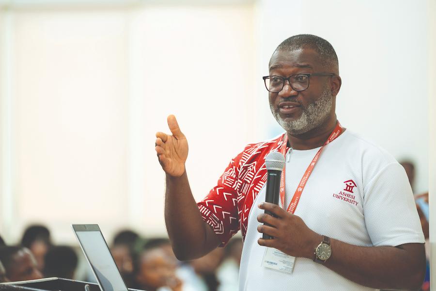 Abdul Mahdi, diretor de assuntos estudantis e comunitários, fala a um grupo de estudantes. A foto mostra um homem negro, vestindo camisa vermelha e branco com o emblema da universidade, segurando um microfone enquanto discursa. Há figuras desfocadas de estudantes ao fundo