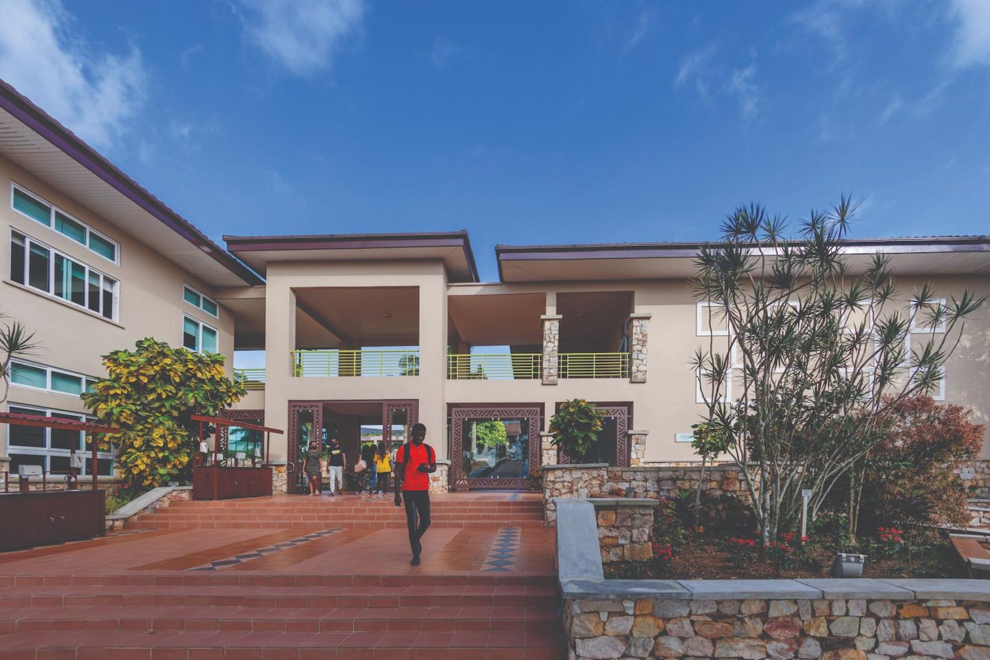 Um estudante atravessa a Founders’ Plaza, na área central do campus da Universidade Ashesi, em Gana. Ele desce escadas feitas de piso marrom, com canteiros de árvores de ambos os lados. Ao fundo, é possível ver um prédio bege de dois andares, com estudantes reunidos no térreo.