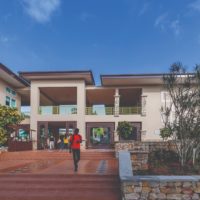 Um estudante atravessa a Founders’ Plaza, na área central do campus da Universidade Ashesi, em Gana. Ele desce escadas feitas de piso marrom, com canteiros de árvores de ambos os lados. Ao fundo, é possível ver um prédio bege de dois andares, com estudantes reunidos no térreo.