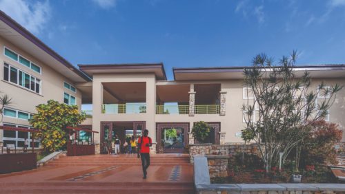 Um estudante atravessa a Founders’ Plaza, na área central do campus da Universidade Ashesi, em Gana. Ele desce escadas feitas de piso marrom, com canteiros de árvores de ambos os lados. Ao fundo, é possível ver um prédio bege de dois andares, com estudantes reunidos no térreo.