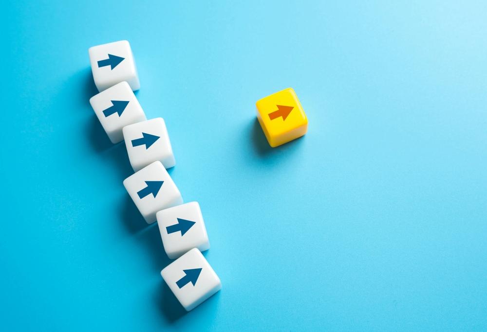 Foto de pequenos blocos com estampas de setas apontadas para frente. Um bloco com uma seta amarela encontra-se mais à frente, enquanto blocos com setas azuis encontram-se atrás, apontando para a mesma direção. A foto ilustra uma posição de liderança