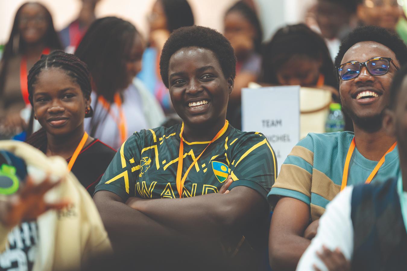 Estudantes da universidade sorriem em primeiro plano. São três jovens negros com blusa verde com detalhes laranja, sendo dois homens e uma mulher. Há outros estudantes ao fundo, fora de foco